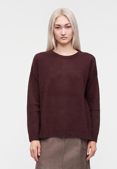 Personne aux longs cheveux blonds portant un pull marron foncé et une jupe grise, debout devant un fond clair uni.
