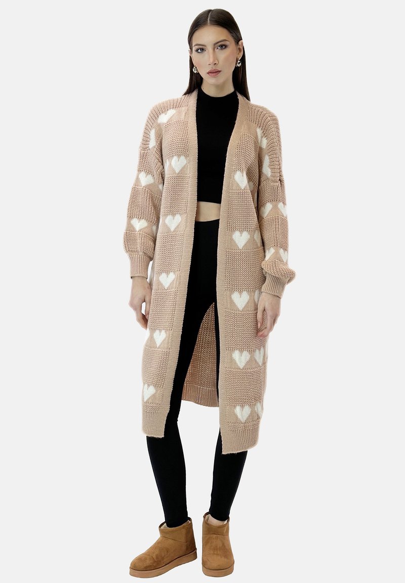 Sweter w kolorze miękkiego beżu z białymi sercami, rozpinany z przodu, z długimi rękawami i ściągaczami, w zestawie z czarnymi legginsami i beżowymi botkami.
