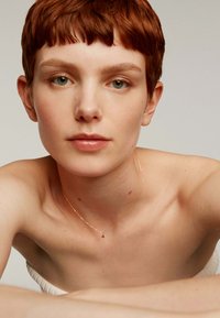 Colgante de estrella dorada en una cadena delgada, lucido contra la piel desnuda. El modelo tiene el cabello corto y texturizado de color rojizo y un maquillaje natural, con un fondo neutro.