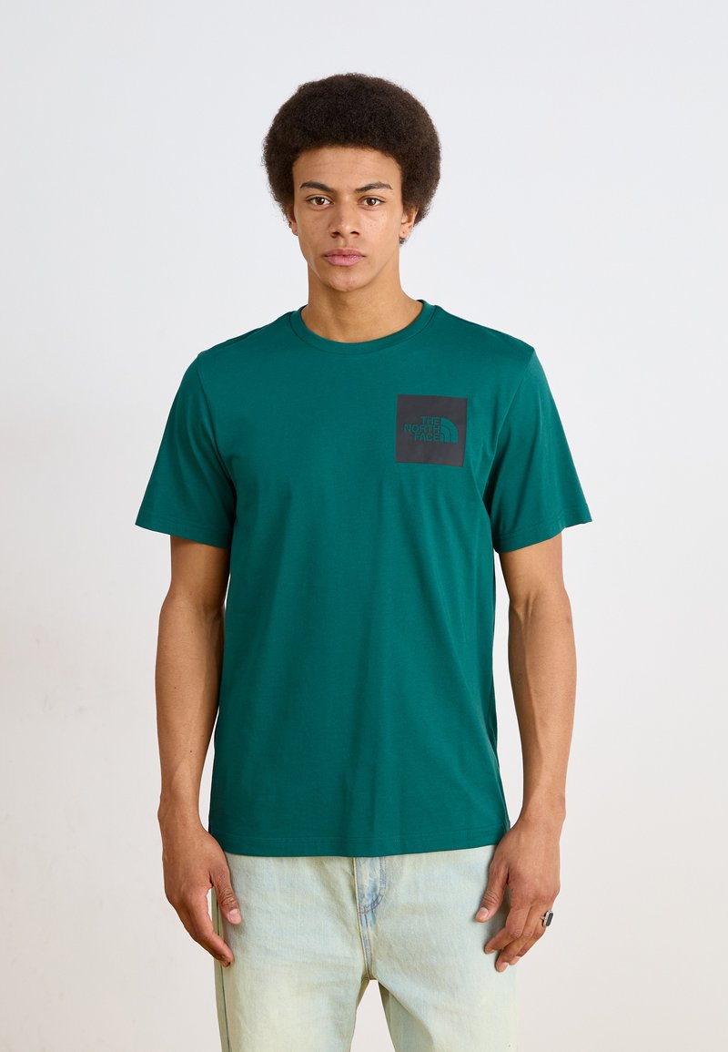 Mlad muškarac s afro frizurom u zelenoj majici The North Face i svijetloplavim trapericama, stoji naslonjen na jednoličnu svijetlu pozadinu.