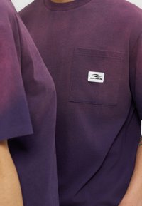 Δύο άτομα φορούν μπλουζάκια με διαβάθμιση μοβ χρώματος, το ένα με τσέπη μπροστά που έχει ένα λευκό ορθογώνιο λογότυπο URTAN.