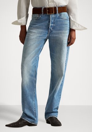 Personne portant un jean droit bleu clair, une ceinture en daim marron, une chemise blanche manches longues et des chaussures noires pointues, debout devant un fond uni.