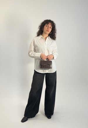 Vrouw met krullend haar, gekleed in een wit overhemd en wijde zwarte broek, die een kleine donkerbruine handtas vasthoudt voor een effen achtergrond.