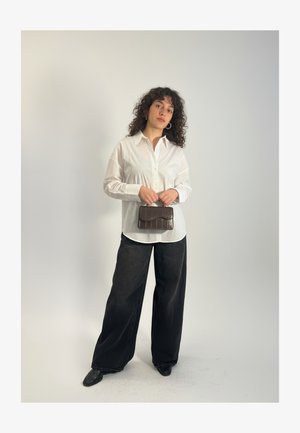 Kobieta o kręconych włosach, ubrana w białą koszulę na guziki oraz szerokie czarne spodnie, trzymająca małą ciemnobrązową torebkę na tle jednolitego tła.