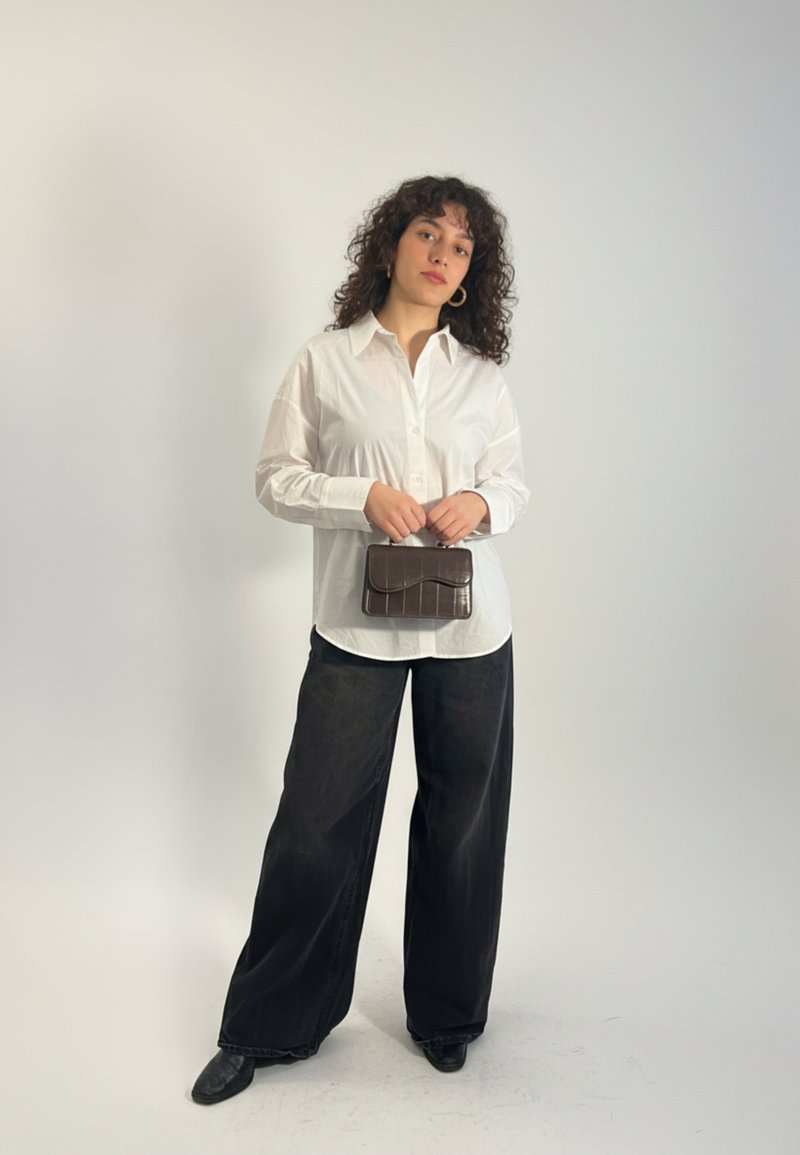 Femme aux cheveux bouclés portant une chemise blanche boutonnée et un pantalon large noir, tenant un petit sac à main marron foncé devant un fond uni.