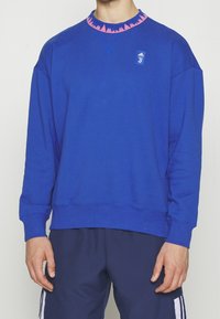Personne portant un sweat bleu Adidas Juventus avec un col rose et un pantalon de sport bleu marine.