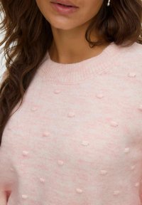 Ženska z dolgimi rjavimi lasmi, oblečena v svetlo roza pleteno pulover s teksturnimi pikami in uhani s perlami, ustnice delno vidne.