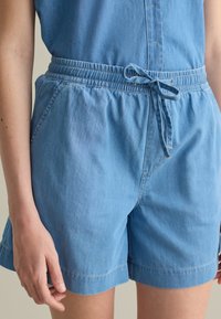 Pantaloni corti in chambray blu con cintura elastica e laccetto. Presentano tasche laterali e una vestibilità rilassata. Tessuto morbido e leggero.