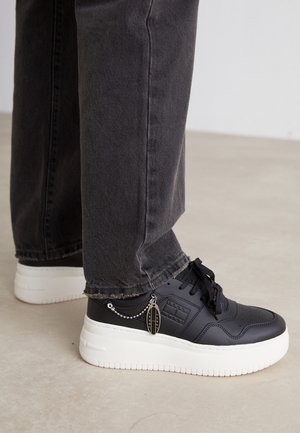 Czarne sneakersy na platformie z białymi podeszwami i małą metalową plakietką z boku, zestawione z ciemnoszarymi jeansami z denimu z postrzępionym dołem.