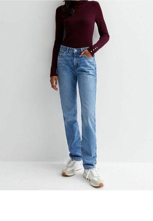Femme portant un jean droit bleu clair, un haut à manches longues bordeaux côtelé, des baskets blanches, debout devant un fond gris uni.