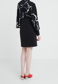 Svart klänning med en åtsittande silhuett och nederkant ovanför knäet, kombinerad med en långärmad blus med ett vitt abstrakt mönster och röda klackskor.