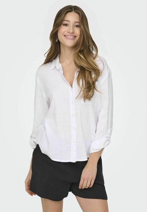 Camicia bianca in lino con colletto, con una vestibilità rilassata e maniche arrotolate, abbinata a pantaloni corti neri. Tessuto morbido, design minimal.