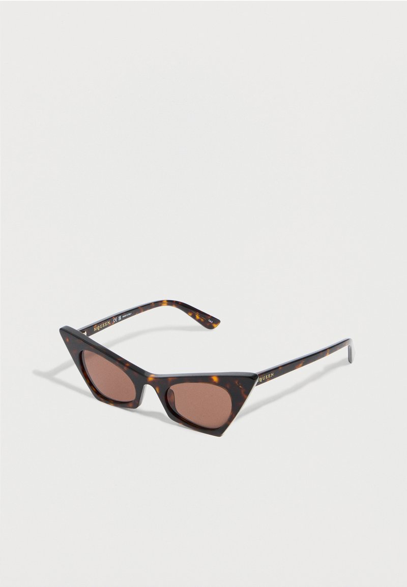 Τριγωνικά γυαλιά ηλίου σχήματος cat-eye με ταρταρούγα, με σκούρους καφέ φακούς και λεπτούς βραχίονες, εκτεθειμένα σε λευκή επιφάνεια.