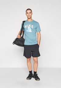 Blå Nike t-shirt med hvide og lyseblå grafik, parret med sorte shorts. Modellen bærer en sort sportstaske og har sorte sneakers på.