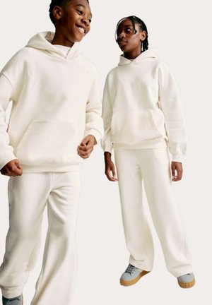 Twee modellen dragen bijpassende crèmekleurige, oversized hoodies met voorzakken en loszittende broeken. Zachte textuur en relaxte pasvorm.
