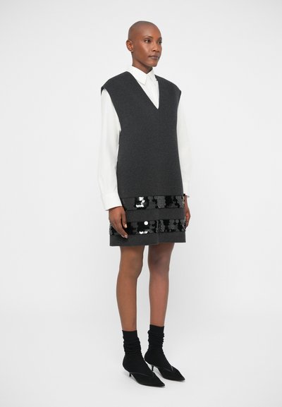 Vestido de suéter cinza sem mangas com um decote em V, sobre uma camisa branca com colarinho, apresentando uma saia preta com lantejoulas. Meias pretas e sapatos de bico.