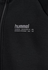 Sort sportstøjsstof med lynet søm og hvid "hummel techwear engineered by HML performance sportswear since 1923" tekst.