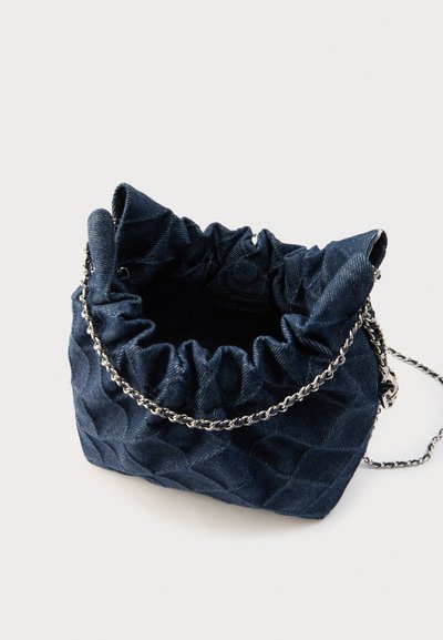 Τσάντα ώμου από σκούρο μπλε βαμβακερό ύφασμα με ραβδώσεις, με ελαφρώς ρυτιδωμένο άνοιγμα και ασημί αλυσίδα ως λουρί ώμου, σε ανοιχτό φόντο.