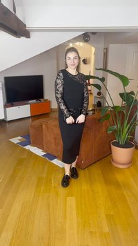 Black midi dress with lace long sleeves, form-fitting design, paired with black shoes. Indoor setting with a plant and furniture in the background.