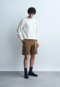 Mann iført hvit langermet skjorte, brune cargoshorts, svarte sokker og svarte loafers, stående mot en ensfarget lys grå vegg.