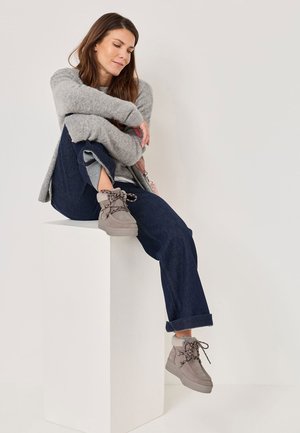 Stivaletti alla caviglia in suede grigio con suole spesse e lacci decorati, indossati con jeans blu e un maglione grigio, seduti su un piedistallo bianco.