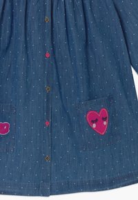 Camisa de mezclilla con un escote fruncido, que presenta pequeños lunares claros, dos bolsillos frontales y parches de corazones rosas con pestañas bordadas.