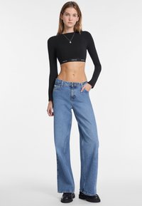 Top negro de manga larga y corto con banda de logo, combinado con vaqueros de pierna ancha en azul claro. La modelo lleva botas negras de plataforma, posando contra un fondo blanco.