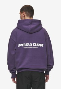 Ljubičasta dukserica s velikim bijelim logom "PEGADOR" i natpisom "CERTIFIED AUTHENTIC STREETWEAR" na leđima. Ima opušteni kroj i kapuljaču.