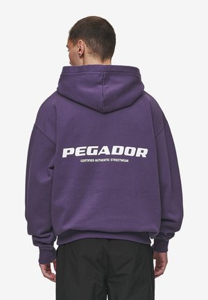 Lila kapucnis pulóver nagy fehér "PEGADOR" logóval és "CERTIFIED AUTHENTIC STREETWEAR" felirattal a hátsó oldalon. Lazább szabású, kapucnival rendelkezik.