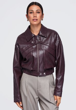 Femme portant une veste en cuir texturée violette foncée avec un large col et un pantalon beige taille haute, debout devant un fond uni.