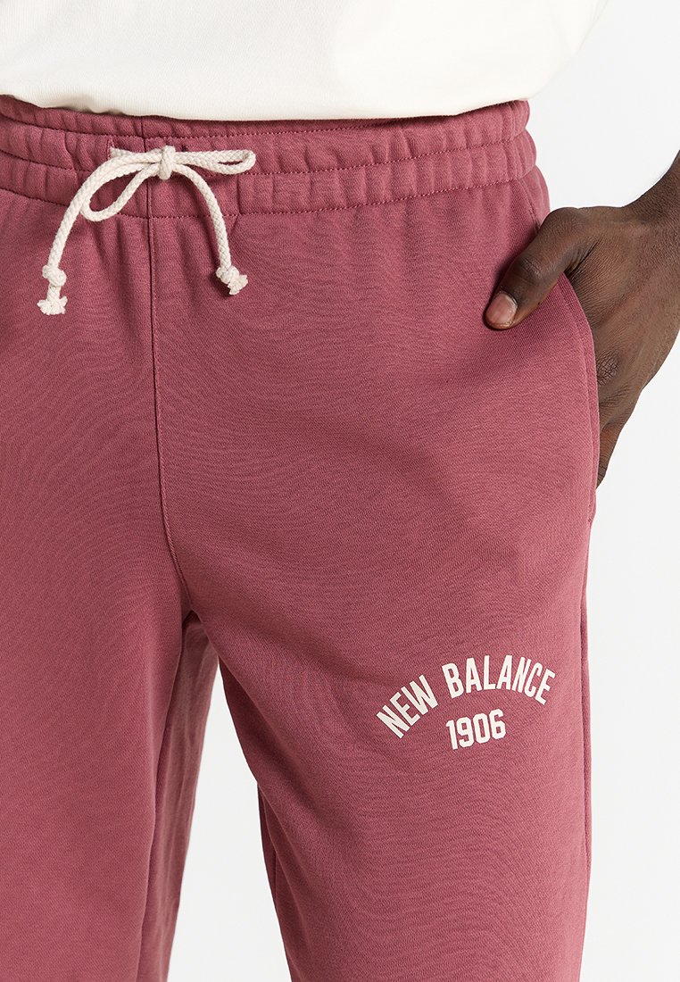 Bordeauxfarbene Jogginghose mit weißem Kordelzug, "New Balance 1906"-Logo auf dem Oberschenkel, getragen mit der Hand in der Tasche, kombiniert mit einem weißen Oberteil.