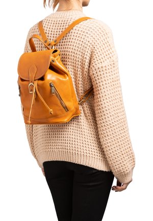 Persoon draagt een beige gebreide trui en zwarte broek met een tan leren rugzak met ritsen en een gesp sluiting.