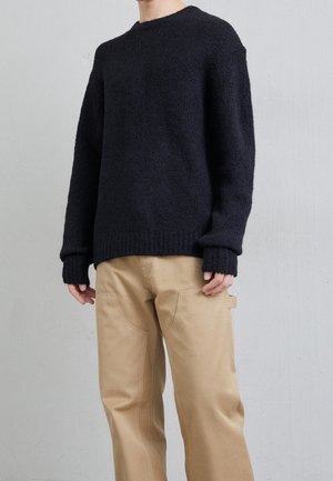 Suéter negro de punto grueso con puños y dobladillo acanalados, combinado con pantalones beiges que tienen bolsillos laterales y detalles de costura decorativa.