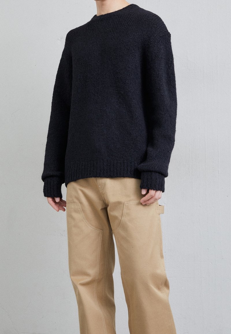 Gruboszyty czarny sweter z prążkowanymi mankietami i dołem, w połączeniu z beżowymi spodniami z bocznymi kieszeniami i dekoracyjnymi detalami szycia.