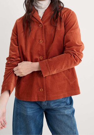 Person, der justerer ærmet på en rust-orange kordjakke med knapper, båret over en hvid rullekrave og blå jeans.