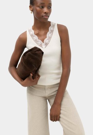 Femme portant un haut sans manches blanc bordé de dentelle et un pantalon beige clair tient un vêtement marron foncé plié devant un fond uni.