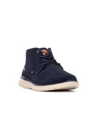 Botín chukka de ante azul marino con punta redondeada, parte delantera con cordones y suela de goma beige; presenta un sutil logo lateral y acentos de cuero en contraste.