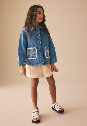 Girl wearing a blue denim jacket with embroidered pockets, beige skirt with floral lace trim, and white sandals standing against a plain background.