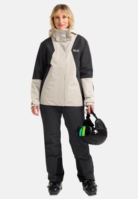 Giacca da sci in grigio chiaro e nero, con colletto alto e tasche. Abbinata a pantaloni da sci neri e un casco con occhialini schermati.