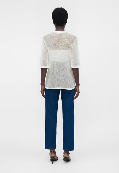 Άτομο που φοράει λευκό τοπ με δίχτυ και μανίκια τριών τετάρτων, μπλε τζιν με ίσια πατζάκια και μαύρα σανδάλια με ψηλά τακούνια, γυρισμένο με την πλάτη.