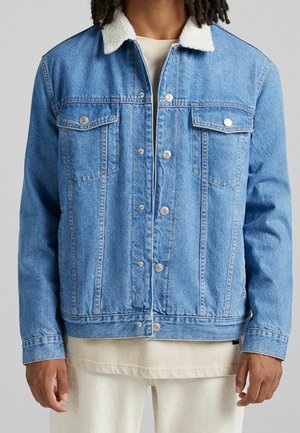 Man draagt lichtblauwe spijkerjack met witte sherpakraag, knoopsluiting, borstzakken en beige broek tegen een effen achtergrond.