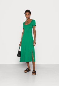 Vestido midi verde de mangas curtas com decote arredondado, fenda lateral e uma silhueta fluida, combinado com sandálias pretas e uma pequena bolsa preta.