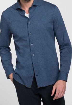 Muž stojící s jednou rukou v kapse, oblečený v modré košili s dlouhým rukávem přes bílé tričko a tmavé kalhoty.