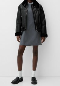 Zwarte leren jas met nepbontkraag, grijze jurk met een gladde textuur, en zwarte loafers gecombineerd met witte sokken.