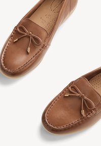 Mocassins en cuir marron avec détails cousus et nœuds décoratifs, présentés sous un léger angle sur fond blanc.