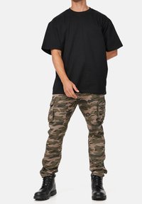 Musta lyhythihainen puuvillapaita yhdistettynä eriparisiin maastokuvioisiin cargo-housuihin, joissa on useita taskuja. Yhdistetty mustiin nauhallisiin saappaisiin.