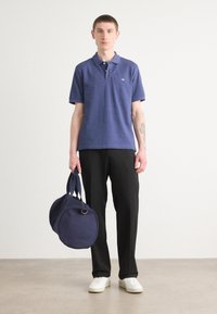 Marineblaues Poloshirt mit kurzen Ärmeln, schwarze Hose und weiße Sneakers. Hält eine runde marineblaue Reisetasche mit blauen Riemen.