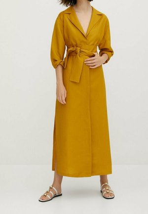Woman wearing mustard yellow long wrap dress with three-quarter sleeves and tied waist, paired with white flat sandals with gold accents.