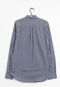 Camisa de cuadros en azul marino y blanco, de manga larga, con cuello, dobladillo curvado, puños abotonados, colgada de un gancho negro.