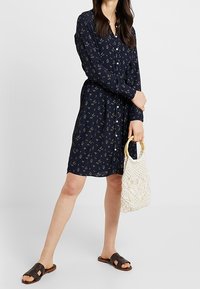 Granatowa sukienka zapinana na guziki z małym kwiatowym wzorem, długimi rękawami i luźnym krojem, w zestawieniu z plecioną torebką i brązowymi klapkami.